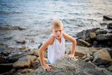 A boy in a white T-shirt and denim shorts peers out from behind a large stone at the seashore.