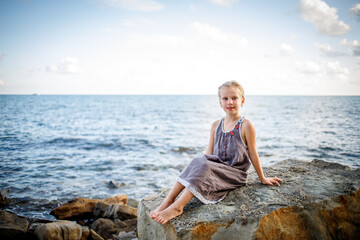 A blonde girl in a sundress sits on a stone on the seashore.