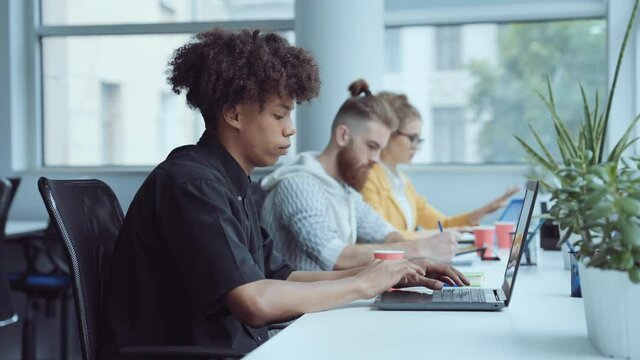 Black man with Afro hairstyle working on laptop in office, diverse coworkers on background. Zoom in employees in shared working environment. Concept of business