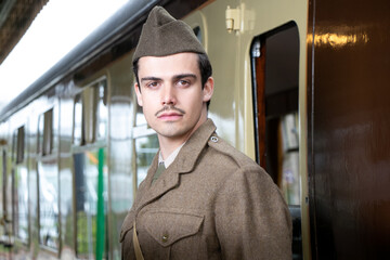 Handsome male British soldier in WW2 vintage uniform at train station next to train