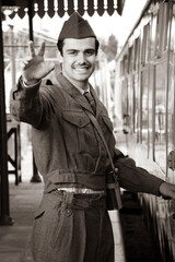 Handsome male British soldier in WW2 vintage uniform at train station next to train, waving and smiling