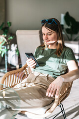 Young pretty woman in green beige clothes sitting in wicker chair and texting in sunlight and shadows, earth colors, using smartphone