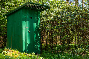 Green timber frame outhouse with heart shape hole in door. Rustic wooden latrine among green vegetation. Dry outside toilet close to non blooming rhododendron bush. Green wood jakes hided among bushes - obrazy, fototapety, plakaty