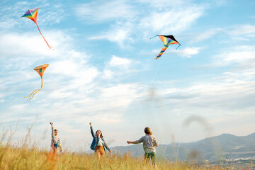 Fototapeta na wymiar Smiling girls and brother boy with flying colorful kites - popular outdoor toy on the high grass mountain meadow. Happy childhood moments or outdoor time spending concept image.