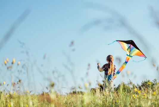 Long-haired Girl with flying a colorful kite on the high grass meadow in the mountain fields. Happy childhood moments or outdoor time spending concept image.