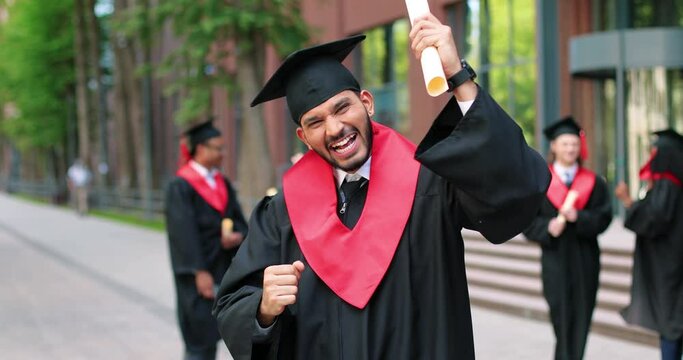 School graduate man in academic gown and hat looking at the camera with happy smile. Happy multiracial man rejoicing near his university or school. Graduation concept