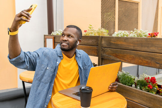 Handsome afro american man in trendy casual wear with yellow laptop in hand sitting in urban cafe outdoors, drinking take away coffee,hold remote talk with business partners or family by phone app