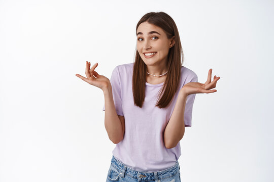 Oops sorry, its me. Cute smiling girl shrugging shoulders and smile awkward, made small mistake, apologizing, dont know, did not understand, standing against white background