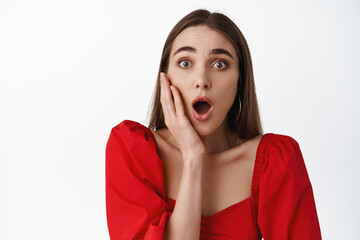 Image of girl in red dress, look amazed and surprised, open mouth and gasp thrilled, hear amazing news, big promo deal, standing in disbelief against white background
