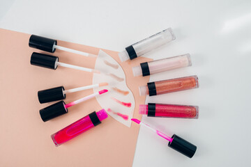 Top view of five opened lip glosses and their brushes placed around white lip-shaped figure and color strocks on it. Makeup color samples.