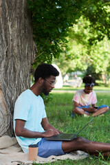 Fototapeta premium African student sitting on the grass and working on laptop in the park