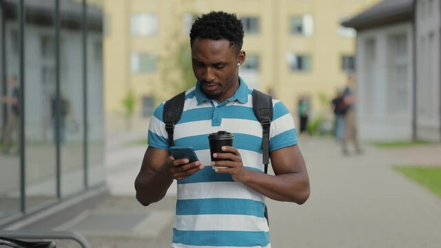 African american man having a walk in the town, enjoying his tasty coffee. Typing messages on his smartphone and drinking hot coffee.