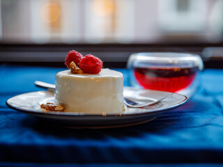 raspberry mousse cake with a glass mug of red tea and a spoon on a white plate on blue textile and open window background, close-up side view, horizontal, copy space.