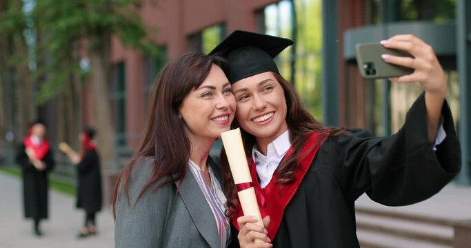 Happy mother and her graduate daughter posing for selfie on graduation day. Girl in academic dress and caps taking self picture with her proud mother