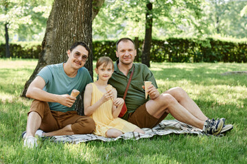Portrait of gay couple sitting on the grass with child and eating ice creams together