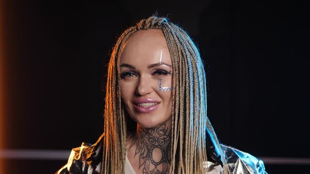 Smiling happiness young adult look. Attractive joyful blonde dreadlocks. Colourful stylish braids. Pink and blue shining. Glowing cheerful face. Perky make up. White face paint and silver jacket 4K.