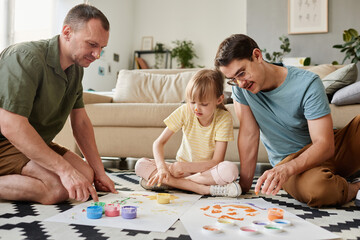 Gay male couple painting on the floor in the room together with their adopted daughter