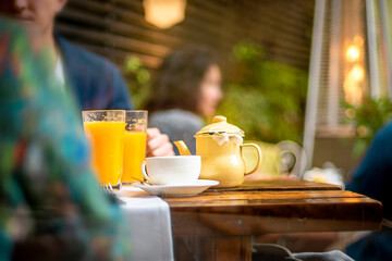 Cafe table for a breakfast with orange juice, tea and coffee. Blurred people sharing food.