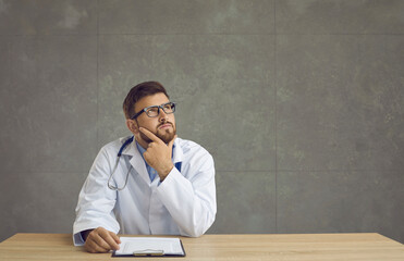 Thoughtful serious millennial doctor rubbing chin while thinking of diagnosis sit at desk with...