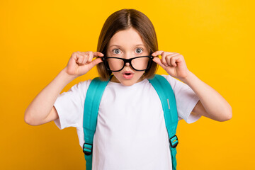 Photo of pretty impressed school girl wear white t-shirt backpack arms glasses big eyes isolated yellow color background