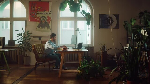 Young Handsome Man Works on a Desktop Computer in Creative Agency in Authentic Loft Office. Renovated Stylish Design with House Plants, Artistic Posters and Big Rounded Windows. Zoom In Handheld Shot.