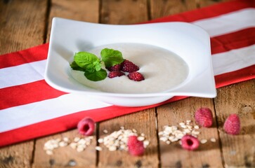 White soup with milk, mint, raspberries and oatmeal on a white plate on wooden background.