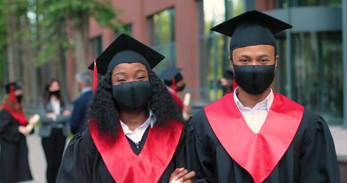 Two happy multiracial student people wearing protective masks are celebrating their graduation while standing near the college and rejoicing with diplomas at the hands. Education concept