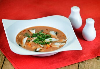 Red, fish soup with papper, olives, parsley and tomatoes on a white plate on wooden background.