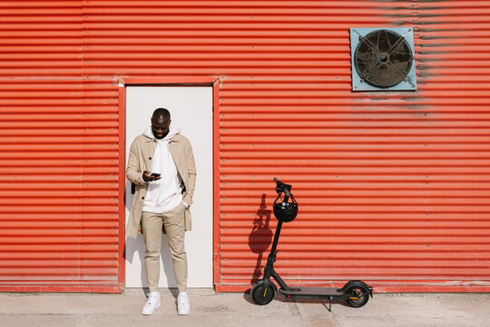 Black man using a mobile in front of a door with electric scooter parked