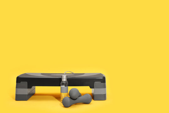 Step platform, dumbbells and bottle of water on yellow background, space for text. Sports equipment