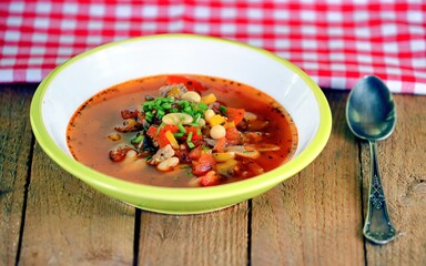 Red vegetable soup with papper, bean, parsley and tomatoes in a white and green bowl on wooden background.