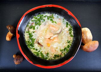 Cream soup with mushrooms, parsley and salmon in a red and black bowl on wooden background.