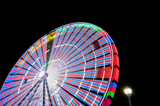 A night time carnival or state county fair ride, an eye or ferris wheel is lit up with neon LED lights as it spins, creation motion trails and sometimes motion sickness for riders aboard