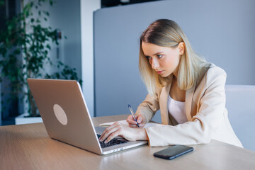 The girl looks at the laptop screen in the office. Serious young woman making notes.