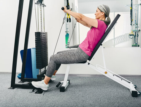 Mature patient sitting on sports bench while using pull cable exercise machine for rehabilitation at rehab center, side view - Powered by Adobe