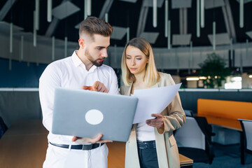 Fototapeta premium Business woman and man with laptop standing and discussing a project in an office building. They look at the computer screen and documents, compare the report.