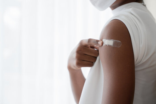 Selective focus on medicated plaster stuck on upper arm of dark skinned man wear mask pointing at plaster. White background. Concept for people to get vaccinated to prevent corona virus.