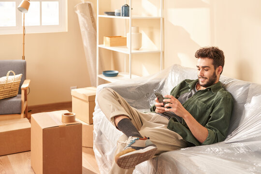 Young man sitting on couch and using cellphone during relocation