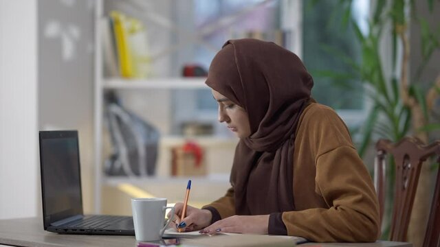 Concentrated confident busy woman writing with pen analyzing e-market on laptop. Side view portrait of focused Middle Eastern analyst in hijab working remotely in home office. Freelance concept