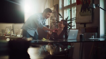 Art Director and Marketing Manager Discussing Work-Related Projects on a Laptop Computer in Creative Agency in Loft Office. Sunny Renovated Space with Plants, Artistic Posters and Rounded Windows.