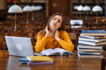 . A young woman student is sitting at a table full of books with textbooks, feeling tired, she is...
