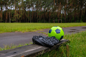 Green soccer ball and soccer boots on the forest stadium background in the morning after the rain. Ukraine.	