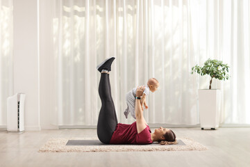 Mother laying on an exercise mat and lifting a baby at home in a living room