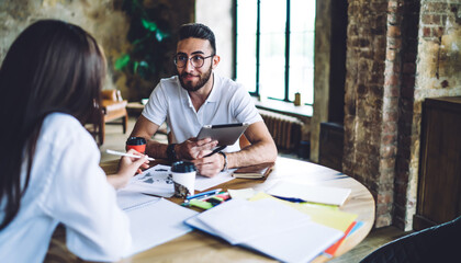 Turkish entrepreneur in optical spectacles for provide eyes correction talking with female colleague for discussing website from touch pad, millennial students learning and studying in coworking
