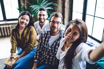 Selfie portrait of cheerful hipster guys smiling at camera while clicking pictures of dream team in loft coworking space, friendly multiracial students enjoying collaboration meeting for communicate