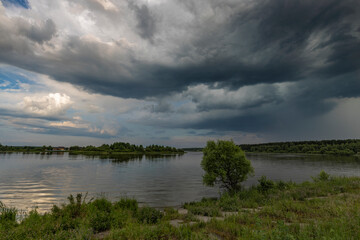 View of the lake from the old dam, overgrown with green grass. The water is brown with slight waves. Thick dark clouds before a storm on an evening summer day.