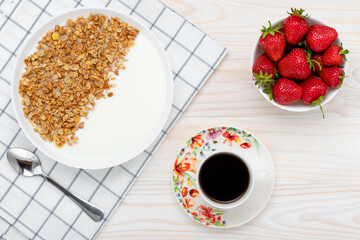 Muesli, a cup of black coffee and strawberry on a wooden table. Hot morning drink. Dry granola with yogurt and red berries, breakfast concept. Healthy vegetarian food. Top view. Flat lay composition.