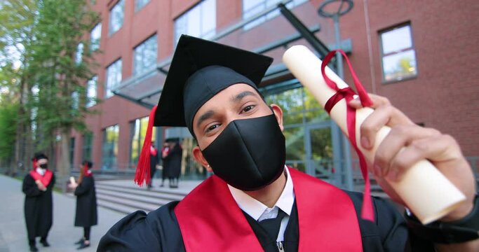 Educational concept. Happy multiracial student boy being happy at his diploma, graduation day. Brunette man standing at the street with his friends at the background and looking at the selfie camera