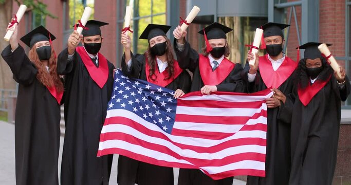 Young graduated people holding their graduation degree convocation ceremony and embracing while posing towards the camera and holding flag of America with background of academy