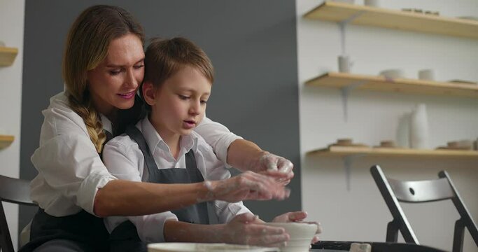 Young mother and her cute baby boy son work together on potter wheel. Handmade ceramic workshop. Slow motion